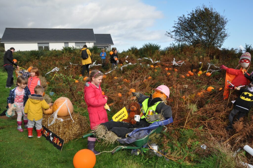 Children select a pumpkin to take home and carve while others admire the scarecrows and carved pumpkins.