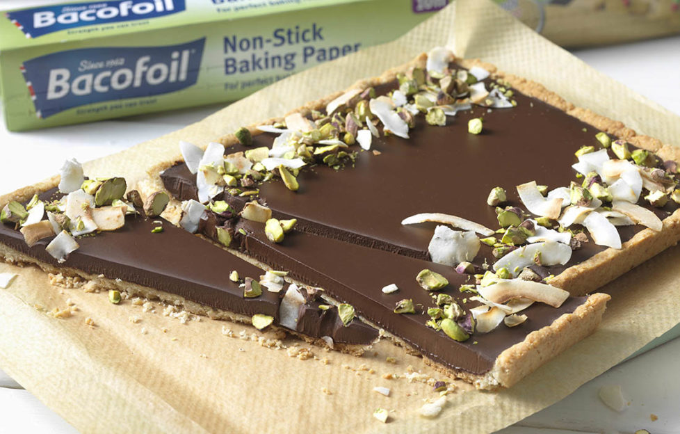 Smooth chocolate tart, decorated with toasted coconut and green pistachios, in baking tray with long, narrow triangular slices cut across the width