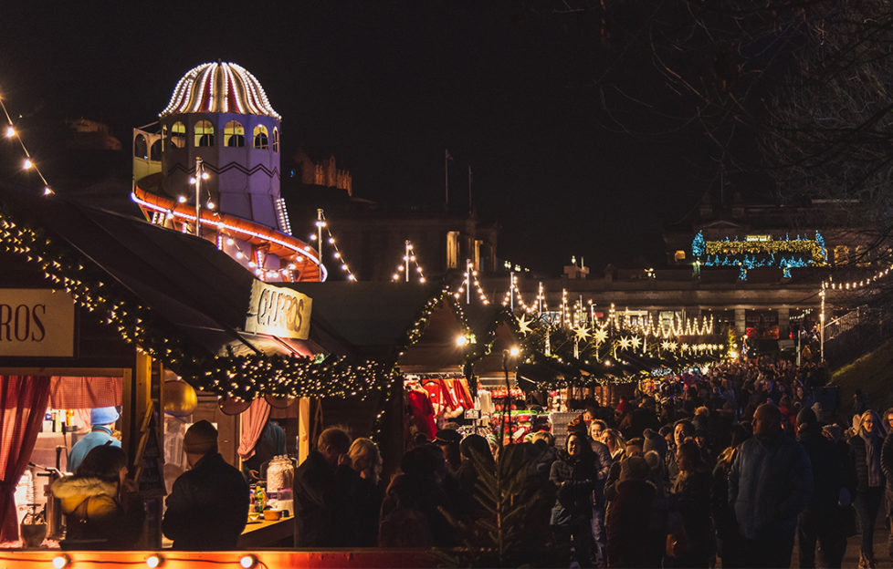 Edinburgh Christmas Market at night with lit wooden cabins, helter skelter and happy crowds