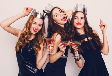 Three young women in black party dresses and silver paper hats, drinking cocktails