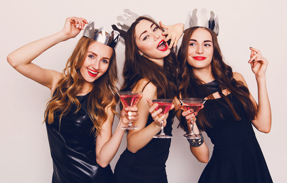 Three young women in black party dresses and silver paper hats, drinking cocktails