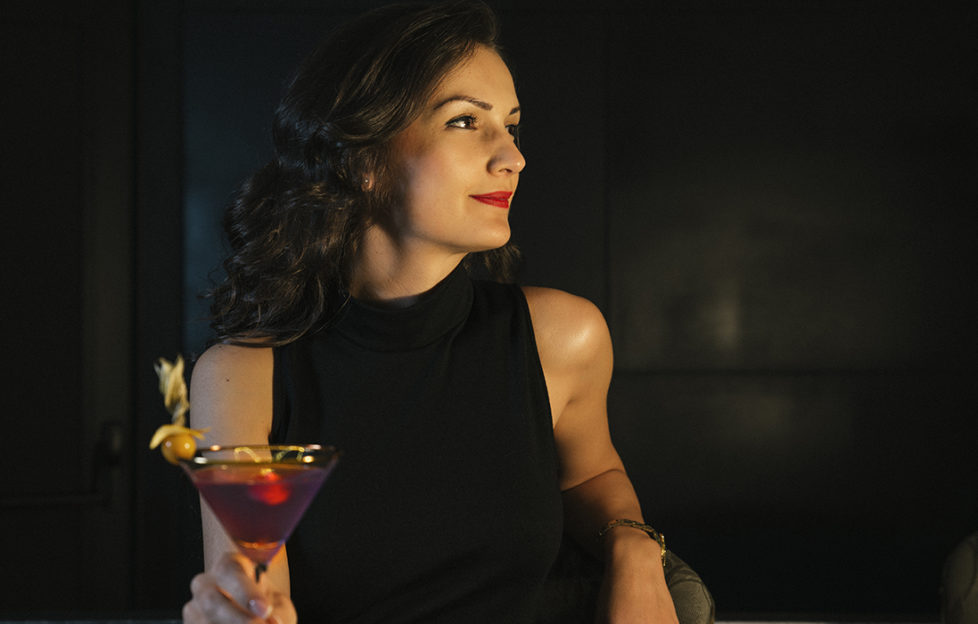 Smiling, relaxed elegant woman in black dress and lipstick, holding red mocktail in conical glass
