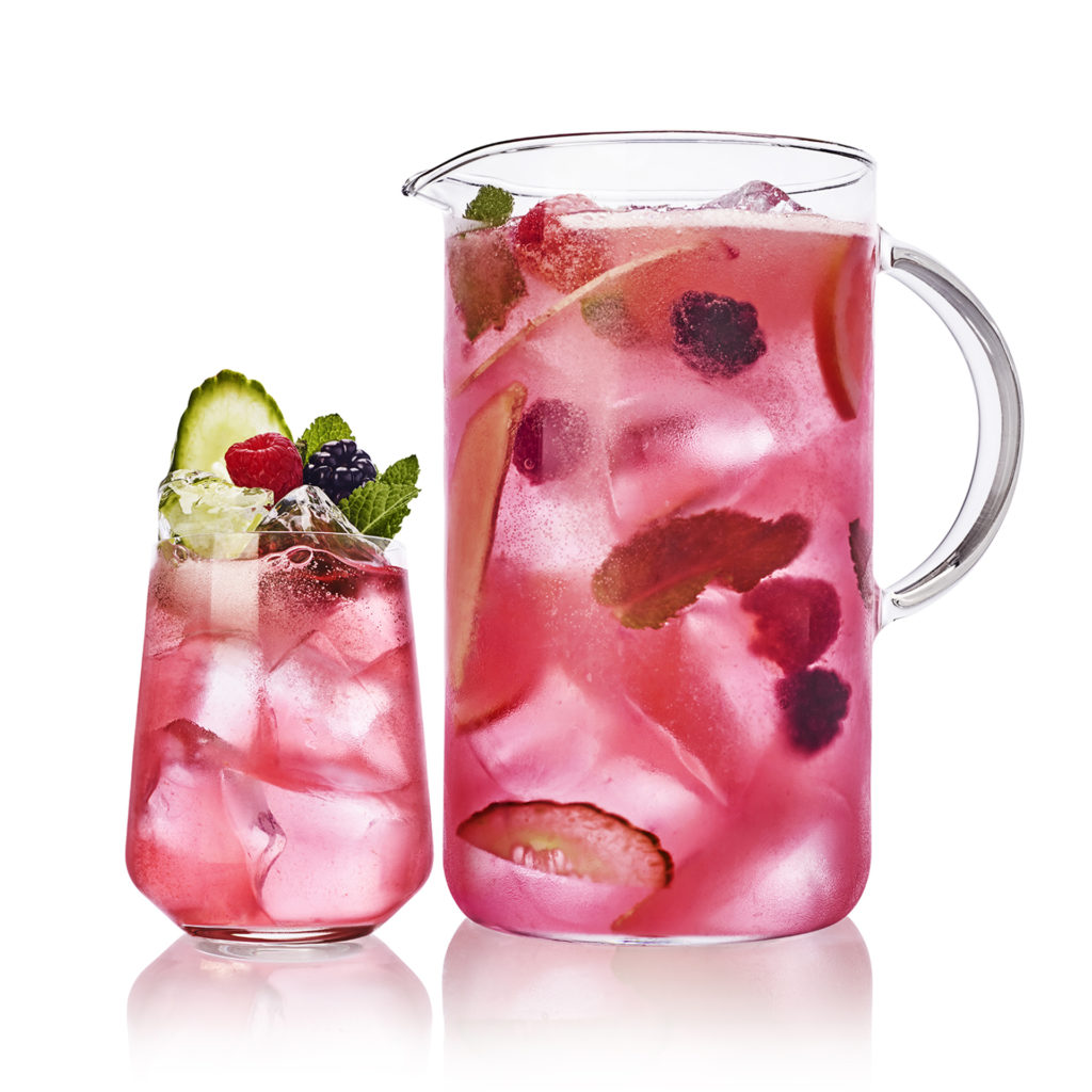 Glass and pitcher of cocktails garnished with cucumber and berries
