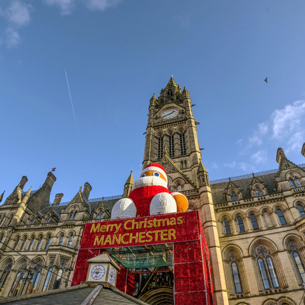 Giant inflatable Santa figure sits on canopy over entrance with message "Merry Christmas Manchester", town hall behind