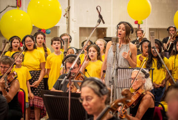 Suranne Jones recording vocal with orchestra members and children's choir in yellow t shirts