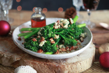 Broccoli, pecans and blue cheese on a serving plate with small glass jar of maple dressing, all on board made of a slice of tree trunk