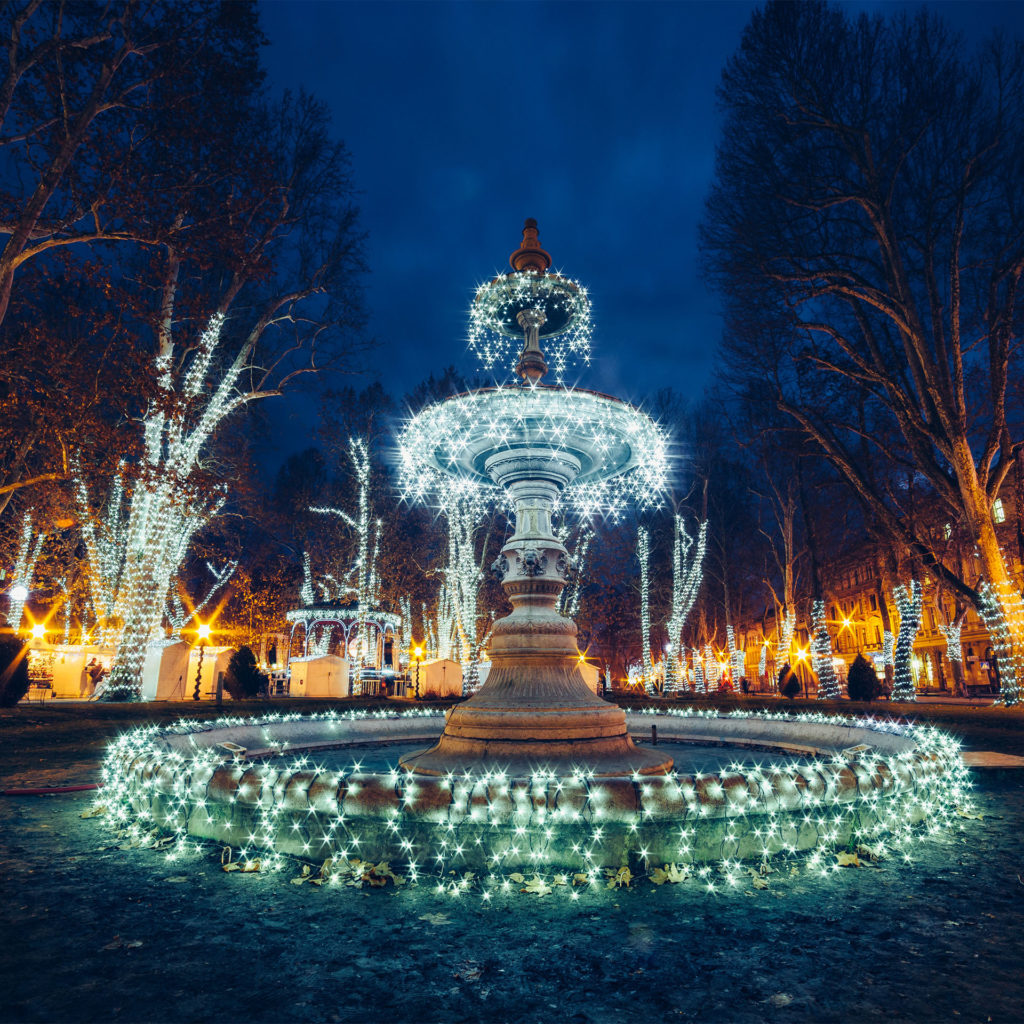 Fairylit fountain in the dark, floodlit classic buildings (same as featured image)