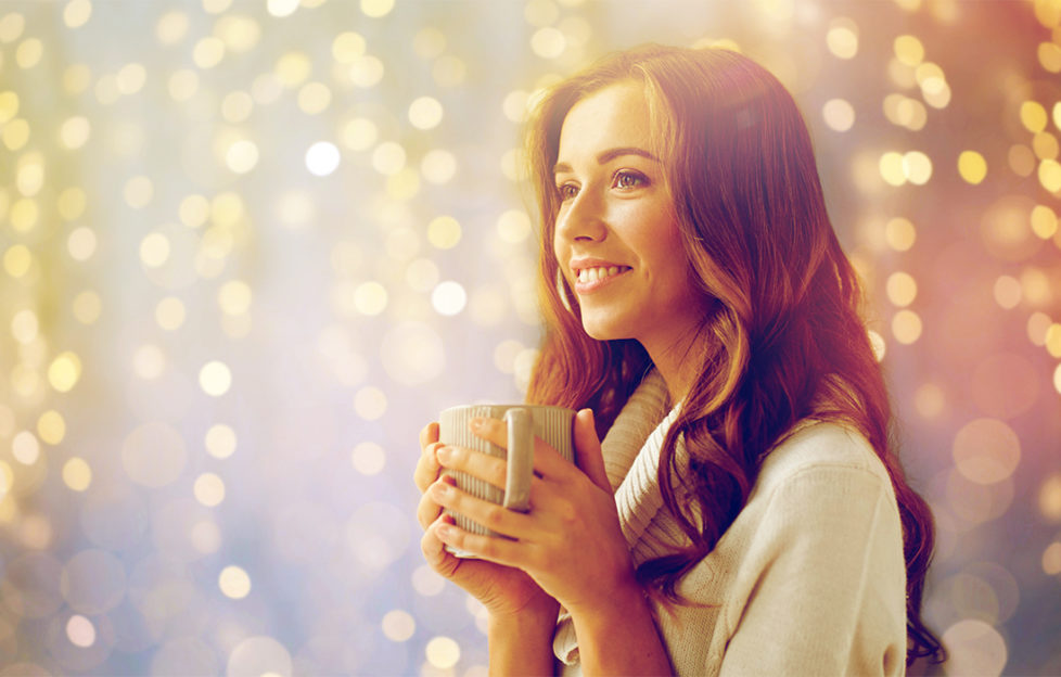 Woman in cosy cream jumper holding mug enjoys atmosphere of soft fairylights