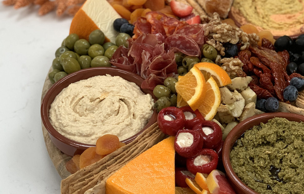 Grazing platter with olives, cured meat, cheese, stuffed peppers, orange slices and dishes of dip,