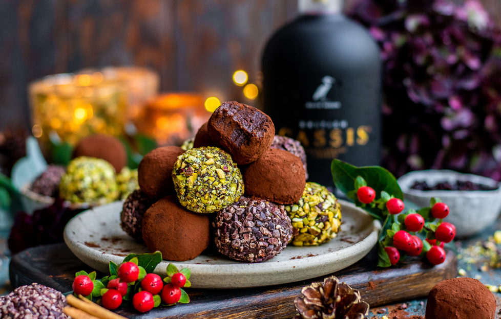 Plte of gorgeous truffles coated in pistachios and cocoa in a pyramid on a plate, one with a bite taken, berries, pine cones and cassis bottle