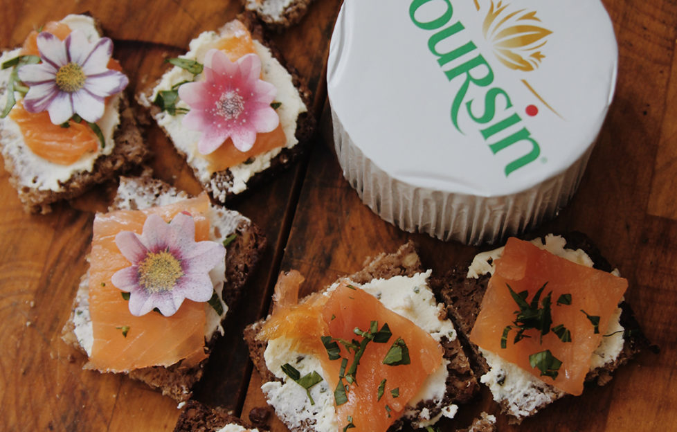 Pack of Boursin soft cheese and 5 small squares topped with soft cheese, smoked salmon, herbs and edible flowers on rustic wooden table