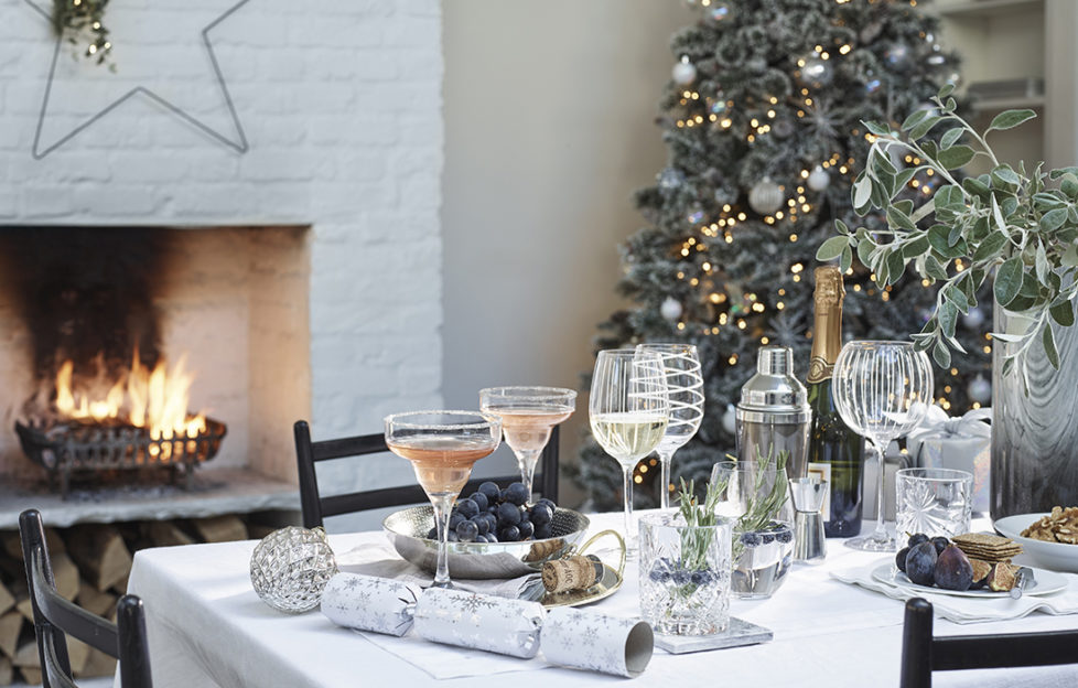 Table set for Christmas dinner with decorated wine glasses and crackers, frost themed Christmas tree behind