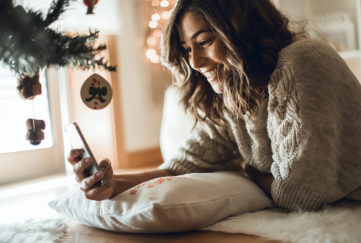 Happy woman using a smartphone lying beside Christmas tree. New Phone? Transfer Your Data YBEC