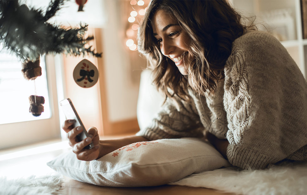 Happy woman using a smartphone lying beside Christmas tree. New Phone? Transfer Your Data YBEC