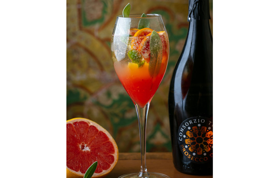 Wineglass of glowing reddish-orange drink with citrus slices and sage leaves