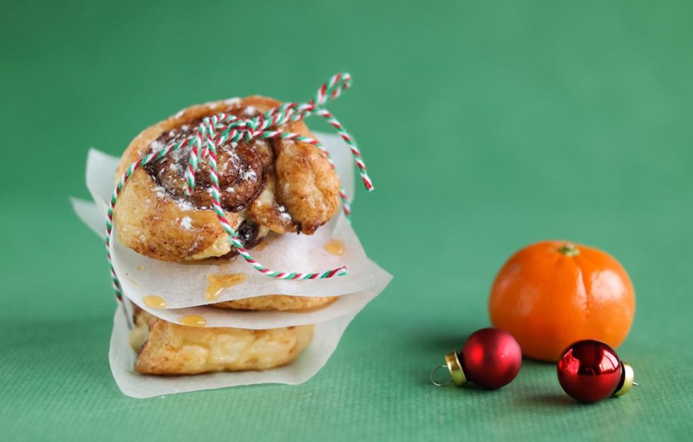 3 cinnamon swirls pastries loosely wrapped in greaseproof paper, tied with red/white/green twisted string, tangerine and baubles on green background