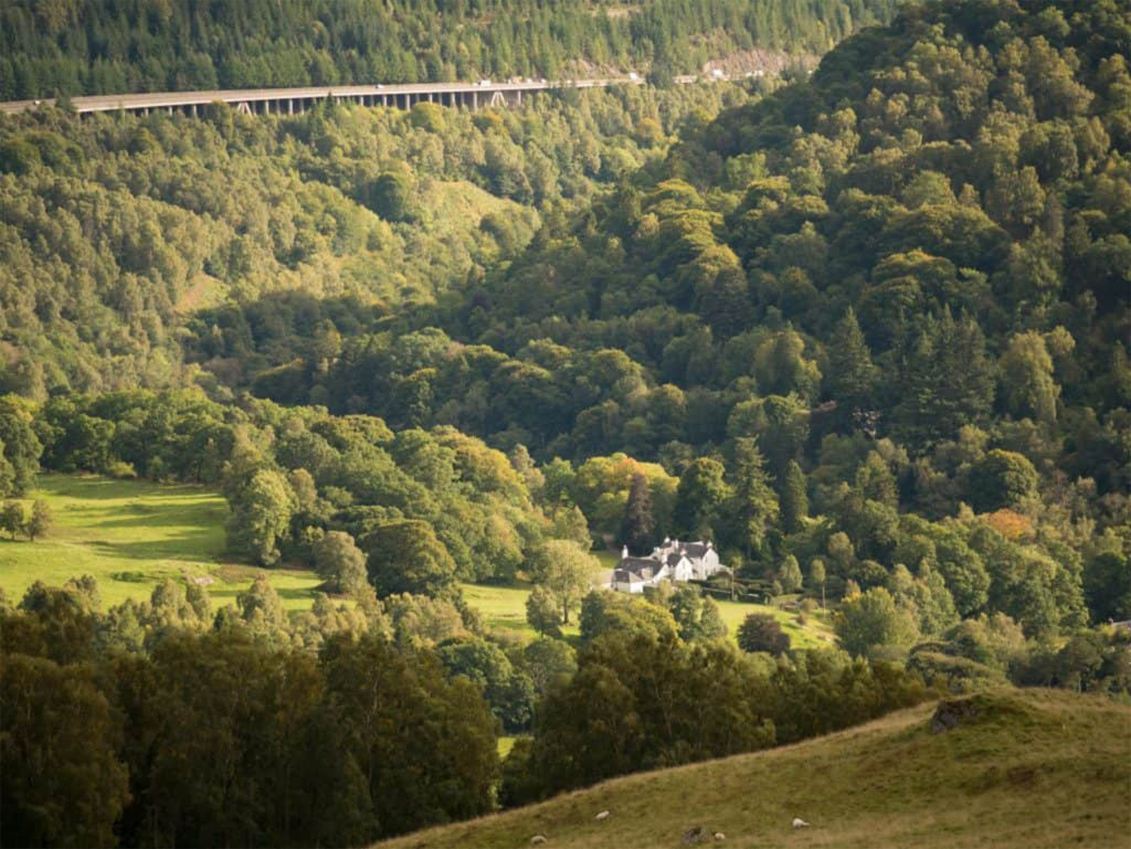 White painted large house almost hidden in wooded hills. Road raised on pillars passes in the distance