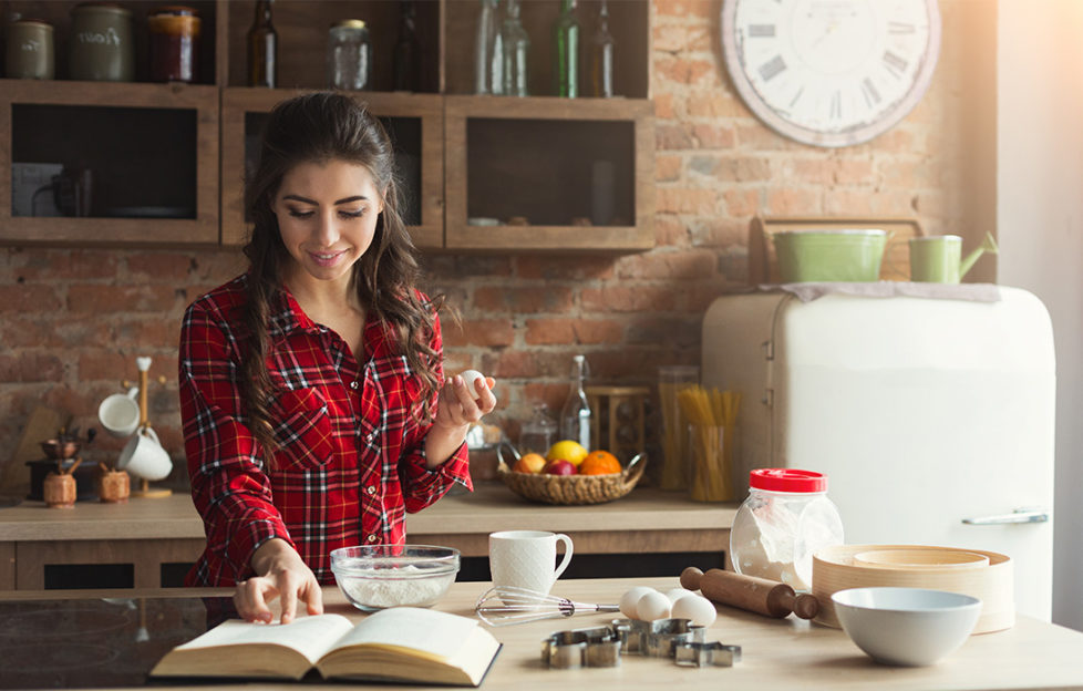 Happy young woman in red check shirt baking pie in loft kitchen at home, using book with recipe