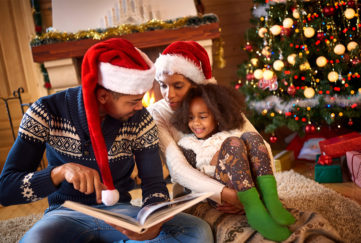 Young parents and girl aged 6 sit by fire and christmas tree reading a storybook together