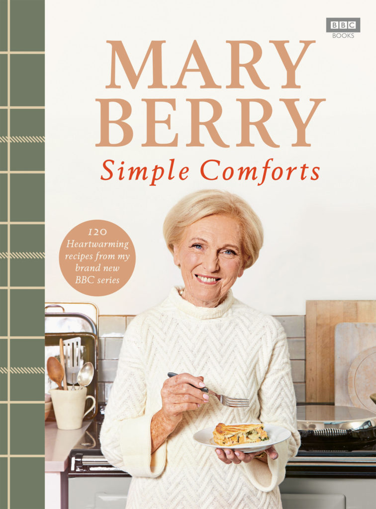 Cover of Mary Berry Simple Comforts, Mary standing in kitchen holding a fork and a plate with a slice of salmon and broccoli pie