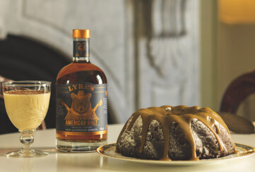 ring shaped fruit cake with caramel sauce, bottle of spirit and glass of eggnog on table