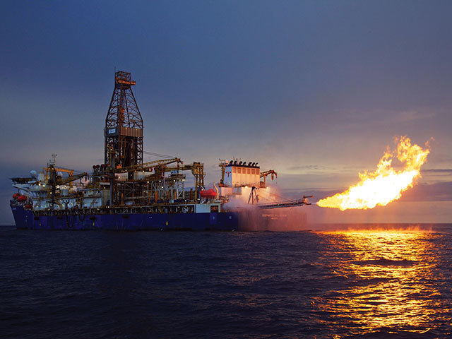 A drillship burns off gas, with the flame reflecting on the azure sea
