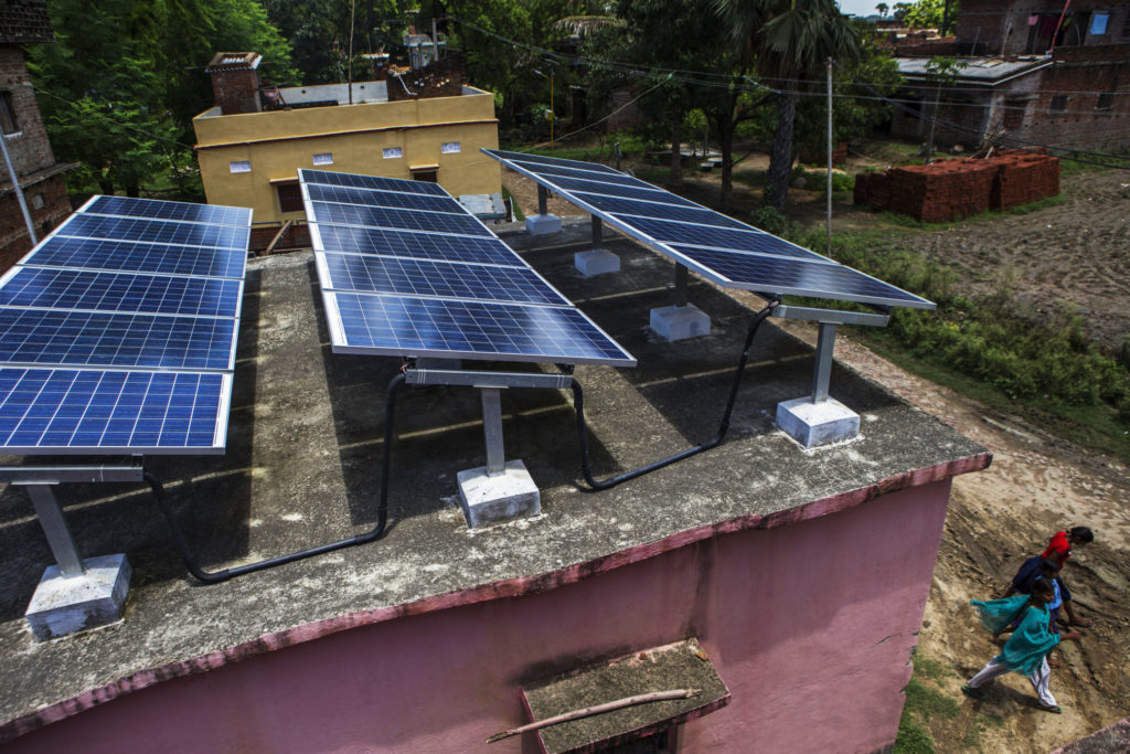 People walk past solar panels mounted on the roof of a building, part of a solar power microgrid, in the village of Dharnai in Jehanabad, Bihar, India. Photographer: Prashanth Vishwanathan/Bloomberg