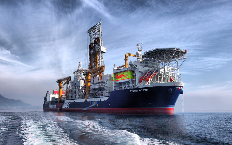 A drillship under a blue sky, with the wake of a boat