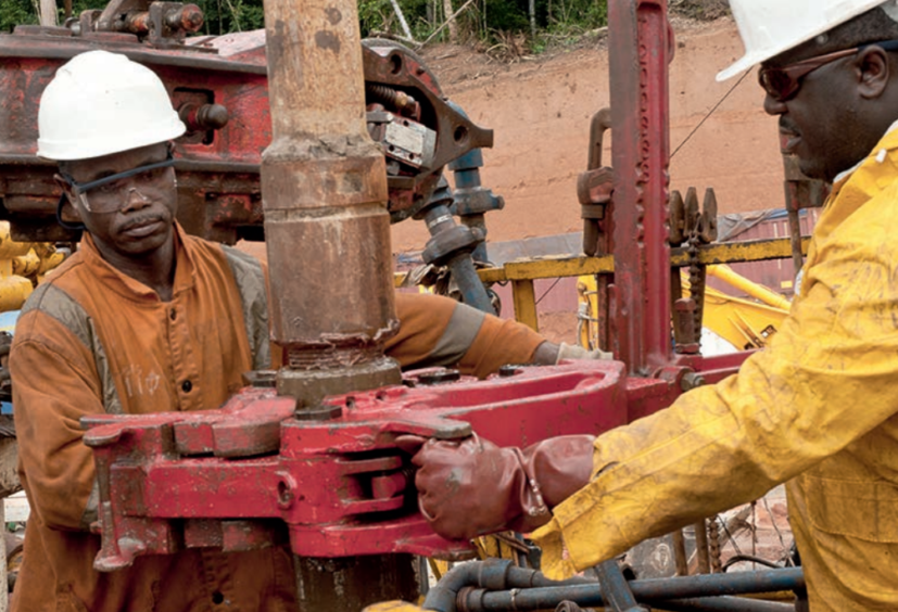 Workers in overalls and hard hats grapple with drilling equipment