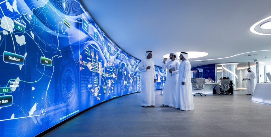 Big computer wall on left, three men talk on the right