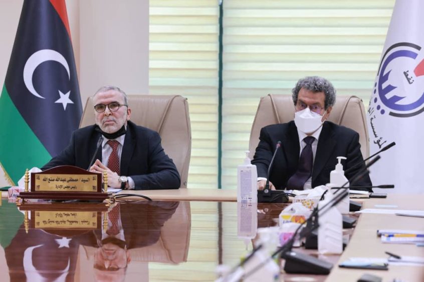 Two men sit at meeting table with Libyan flag