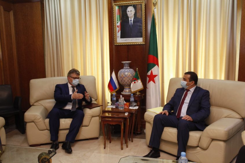 Two men sit and talk with an Algerian flag behind.