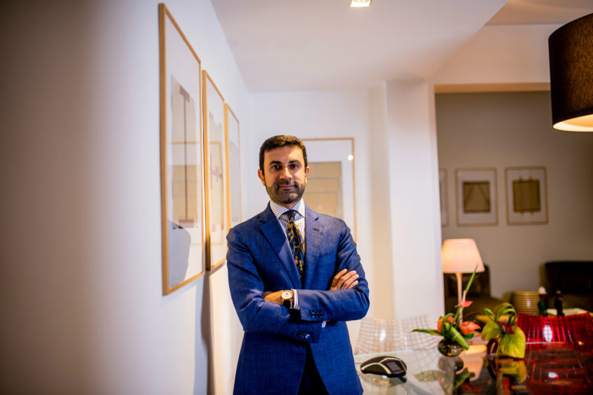 A man in a blue suit with arms folded, a wall on his right