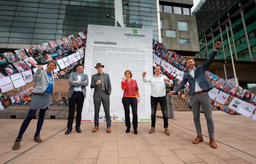 Six people stand on a pavement holding pictures, with a poster behind