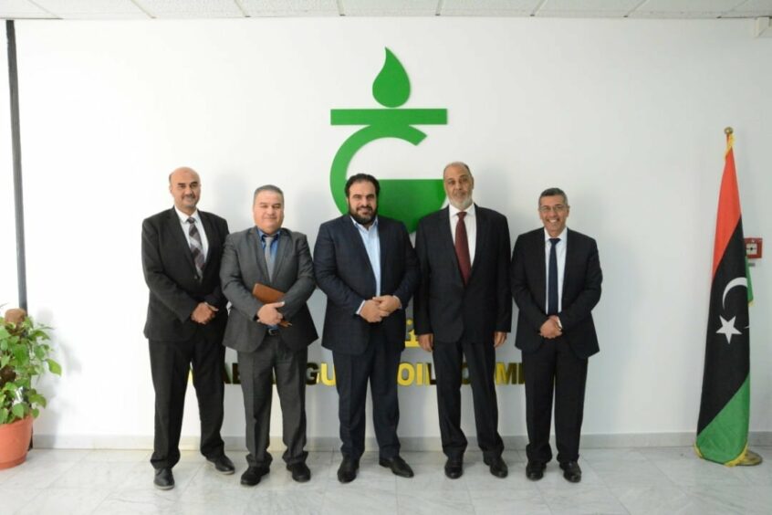 Five men stand in front of giant green logo with Libyan flag to the right
