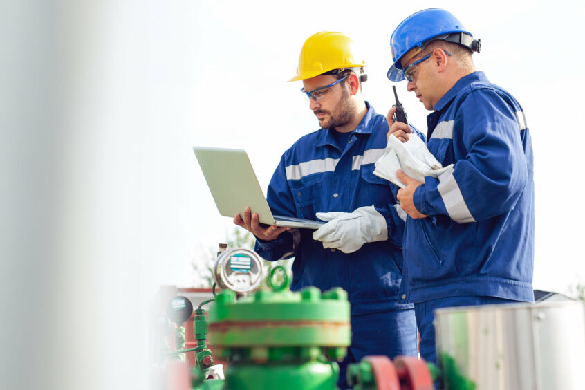 Two men in overalls look at pipe and a laptop