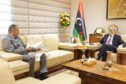 Two men sit on sofas with a Libyan flag behind