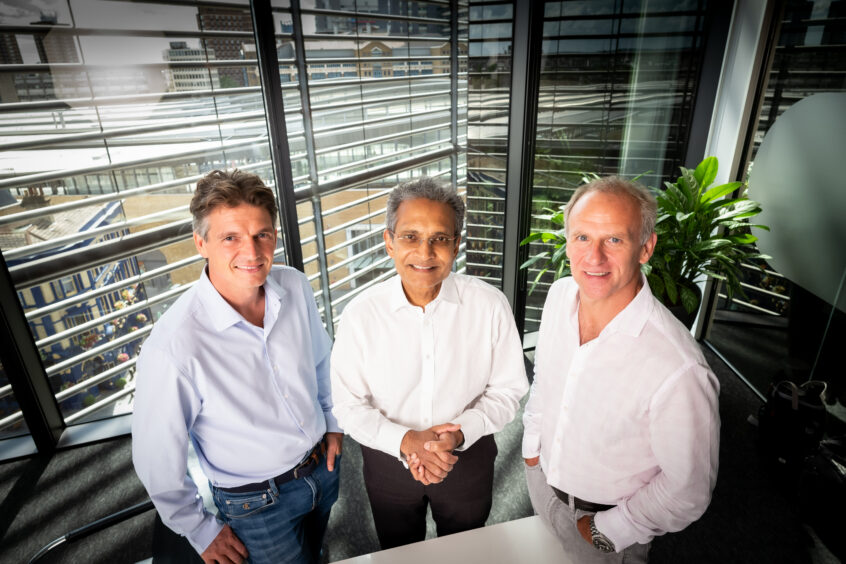 Three men in shirts look at camera, with open blinds behind