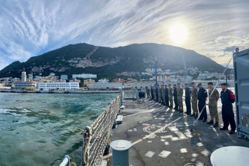 Line of men on a ship's deck on the right hand side, with a mountain behind and water to the left