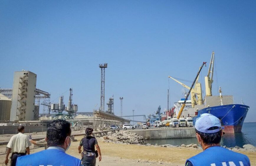 People walking in sunshine at a port with ships in background