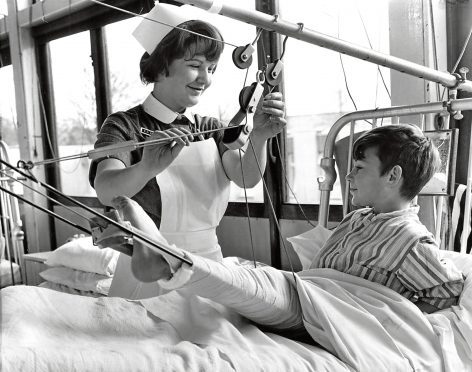 1968: A nurse smiles while she helps a boy with his leg in splints.