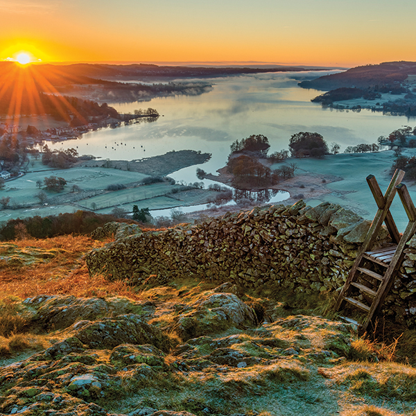Wooden stile and stone wall with Lake Windermere in background with sun rising above horizon Pic: Istockphoto