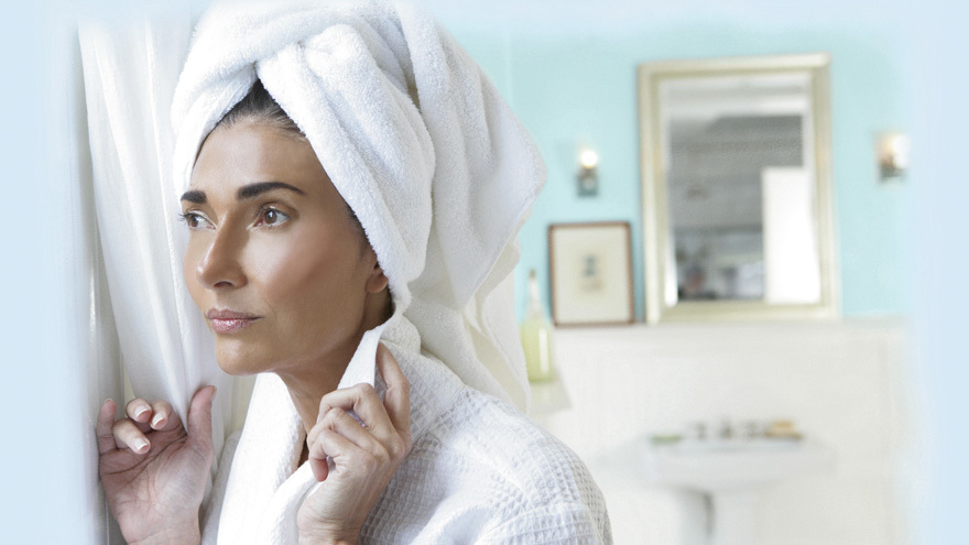 Woman with towel on head looking out bathroom window.