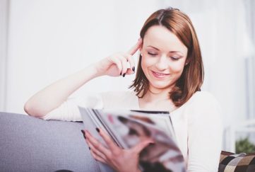 Attractive happy brown haired girl relaxing on couch and reading a magazine