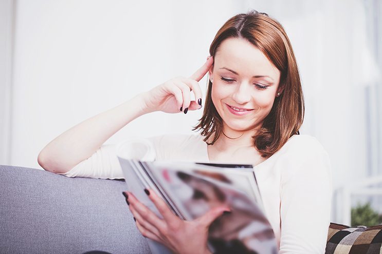 Attractive happy brown haired girl relaxing on couch and reading a magazine