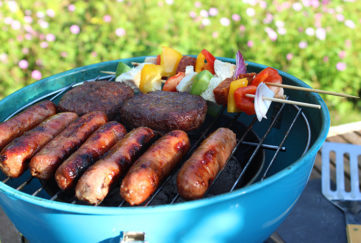 Photo showing a portable kettle barbecue / charcoal BBQ in a garden setting, stood on a wooden patio table with the lawn and pink hardy geraniums forming the blurred background. Pictured cooking on the barbecue are some sausages, beef burgers and colourful kebabs, made from red, green and yellow bell peppers, red onions and chicken pieces.