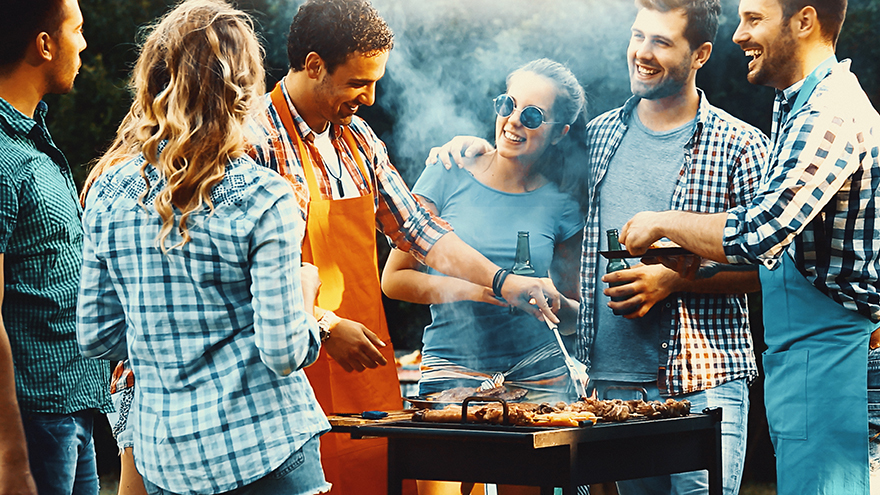 Closeup side view of group of mid 20's people having backyard barbecue party. There are three guys and four girls gathered around heavily smoking grill and sipping cold beer. One of the guys is being today's chef. Toned image, mild contrast, back lit. Pic: Istockphoto