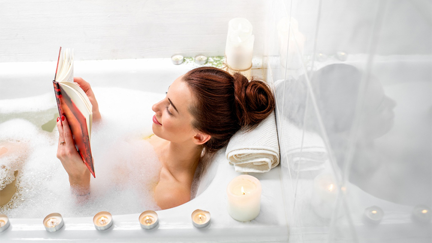 Woman lying in bubble bath reading a book, surrounded by scented candles