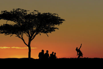 Cover of Listen To The Drums - a tree and a group of people silhouetted against a sunset in a cleat sky