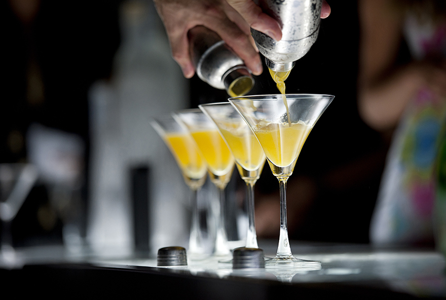 Photograph of human hands holding two cocktail shakers and pouring orange color cocktail into two martini glasses